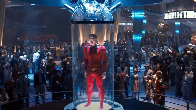 A mannequin in a red spandex suit inside a glass tube is examined by a crowd.