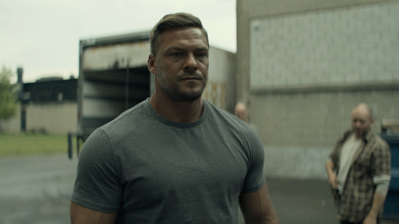 Alan Ritchson's Jack Reacher stands outside surrounded by guards in Reacher