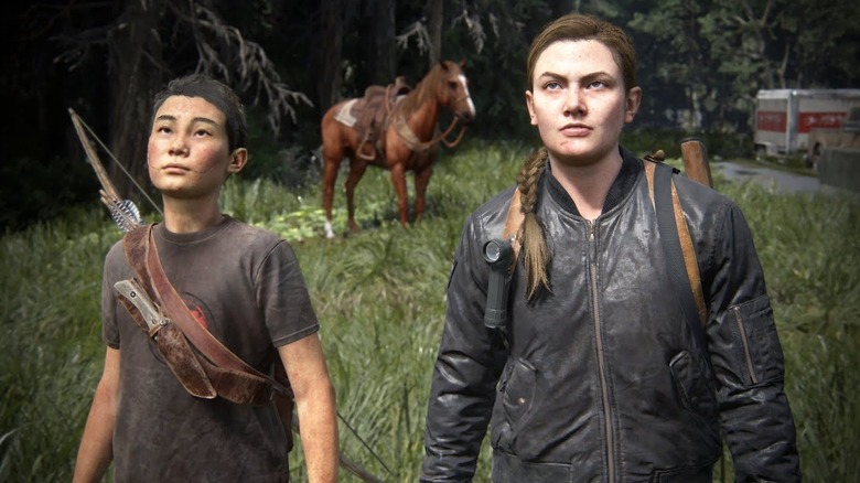 Ian Alexander as the voice of Lev standing in a field and wielding a bow and arrow next to Laura Bailey as the voice of Abby in The Last of Us Part II
