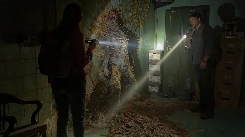 Joel and Tessa point their flashlights at an infected fused to a wall in The Last of Us