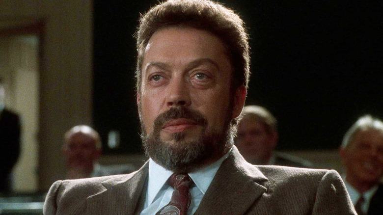 Tim Curry as Herkermer Homolka smiling menacingly in a suit in Congo