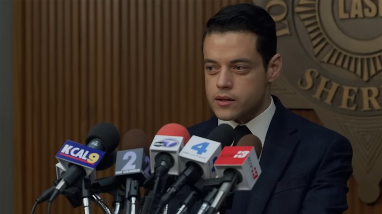 Rami Malek as Baxter in The Little Things, giving a press conference in front of a bunch of microphones