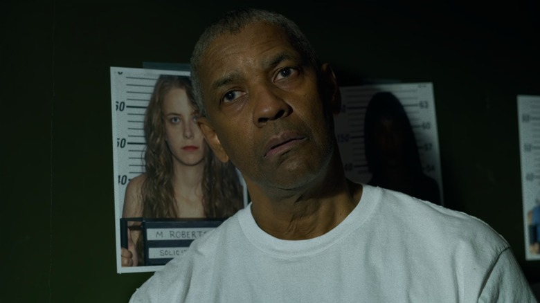 Denzel Washington as Deacon in The Little Things, in a white shirt, head cocked, in front of a wall of mugshots of women
