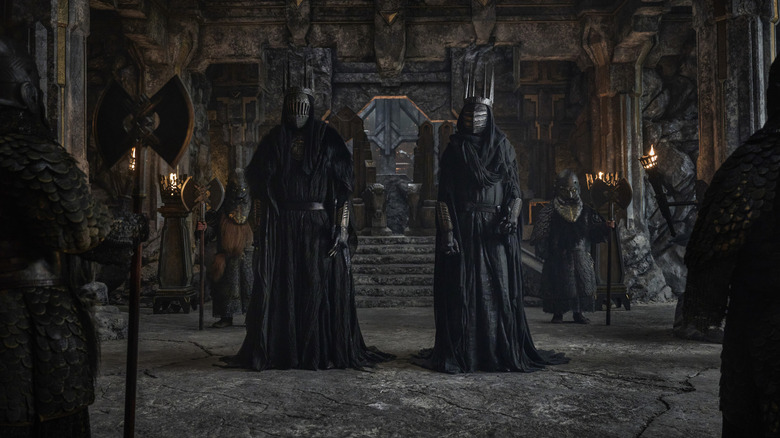 Two robed figures dressed in black and wearing masked helmets standing in a room of stone surrounded by guards with axes in The Lord of the Rings: The Rings of Power