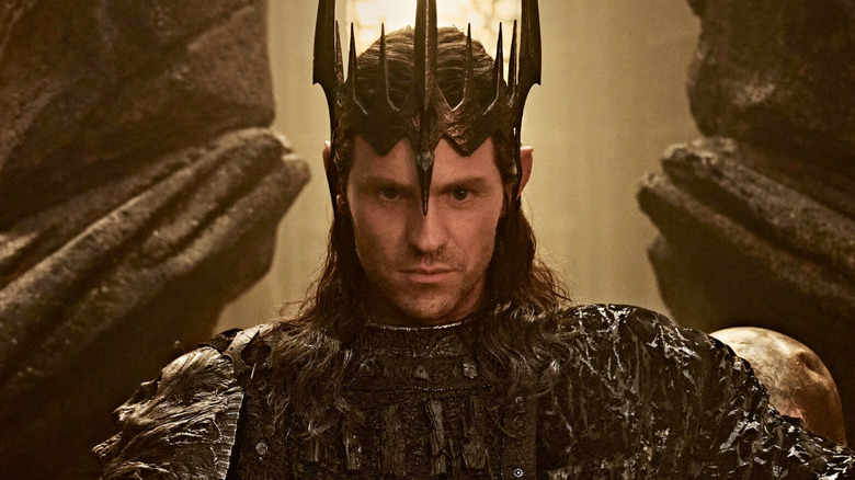 Charlie Vickers as Sauron wearing a crown and seated on a throne in The Lord of the Rings: The Rings of Power