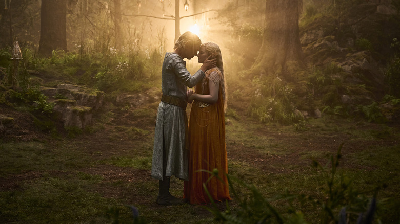Jamie Campbell Bower as Celeborn embracing Morfydd Clark as Galadriel in a sunny forest glade in The Lord of the Rings: The Rings of Power