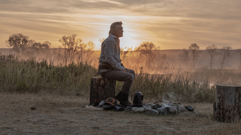 Kurt Russell as Preston Clyburn sitting on a tree stump by a campfire as the sun rises behind him in The Madison