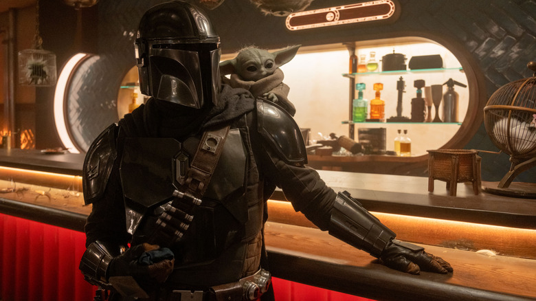 Pedro Pascal as Din Djarin wearing his armor and leaning against a bar with Grogu on his shoulder in Star Wars: The Mandalorian and Grogu