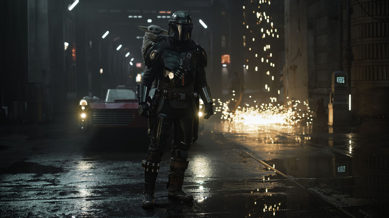 Pedro Pascal as Din Djarin in his armor standing in an empty street with sparks in the background and Grogu on his shoulder in Star Wars: The Mandalorian and Grogu