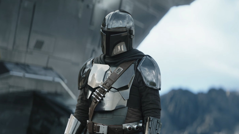 Mando with his head cocked standing by the Razorcrest in The Mandalorian and Grogu
