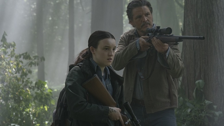 Pedro Pascal as Joel Miller aiming his rifle in a forest next to Bella Ramsey as Ellie in The Last of Us