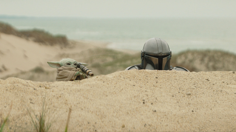 Grogu looks the wrong way through a telescope as he and Mando peer over a sand dune in The Mandalorian and Grogu