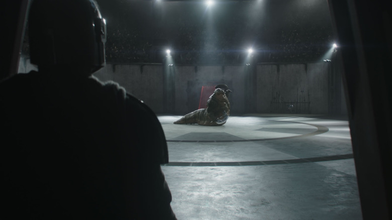 Pedro Pascal as Din Djarin standing by a doorway to an arena watching Jeremy Allen White as the voice of Rotta the Hutt in front of a huge audience in Star Wars: The Mandalorian and Grogu