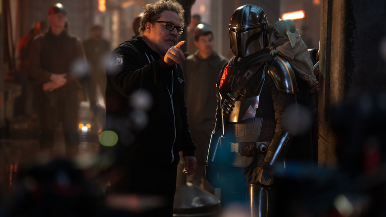 Director Jon Favreau gesturing to Pedro Pascal as Din Djarin on a busy street filled with cast and crew on the set of Star Wars: The Mandalorian and Grogu