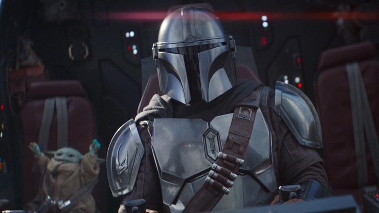 a person in a metal suit sitting in a space ship in the foreground with baby yoda cheering in the background