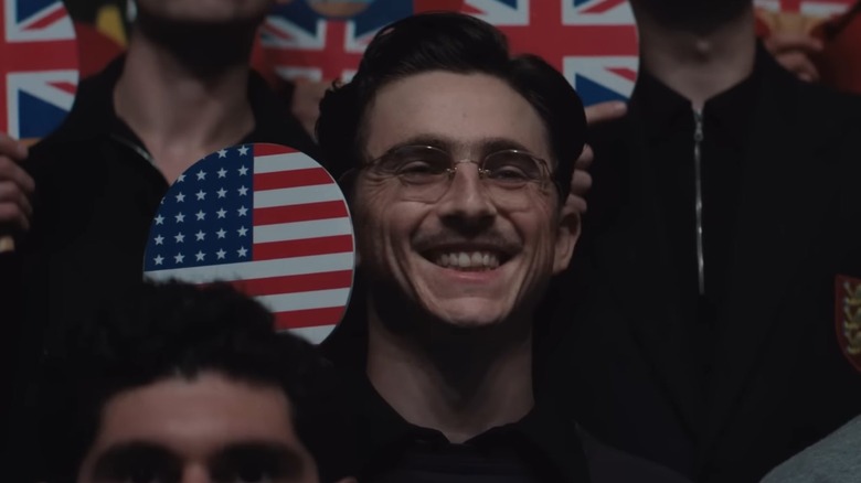 Timothée Chalamet as ping pong star Marty Reisman standing in a crowd and holding up a paddle with the American flag printed on it in Marty Supreme