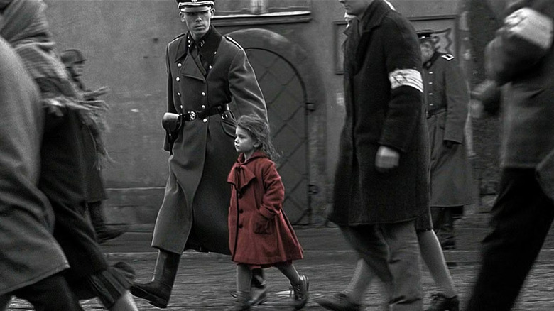Una niña con un abrigo rojo (Oliwia dąbrowska) camina con una expresión concentrada, manos en sus bolsillos, rodeada de hombres judíos con abrigos y brazaletes marcados con estrellas de David, mientras un oficial de las SS los mira mientras camina en la lista de Schindler