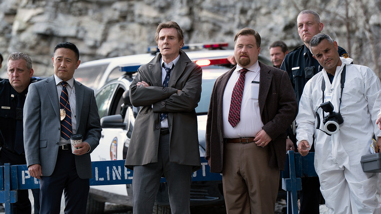 Frank Drebin Jr. (Liam Neeson) and Ed Hocken Jr. (Paul Walter Hauser) and other cops look at a crime scene in The Naked Gun (2025)
