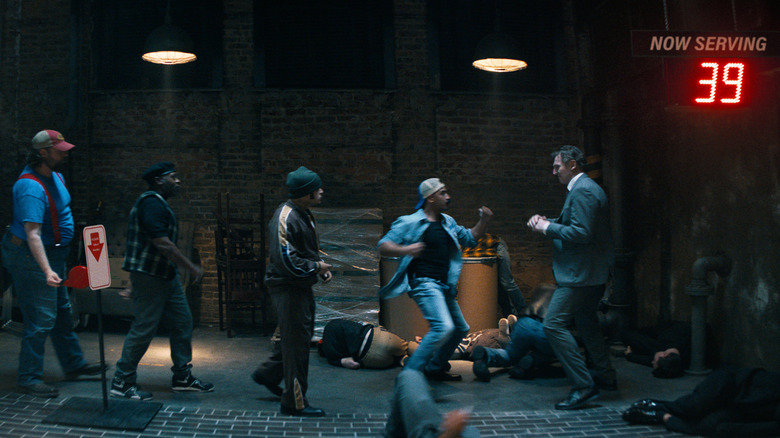 Frank Drebin Jr. (Liam Neeson) fighting a line of criminals with a "Now Serving" number sign above him in The Naked Gun (2025)