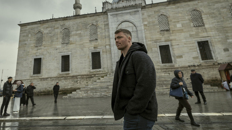 Gabriel Basso as Peter Sutherland walking by a building in the rain in The Night Agent