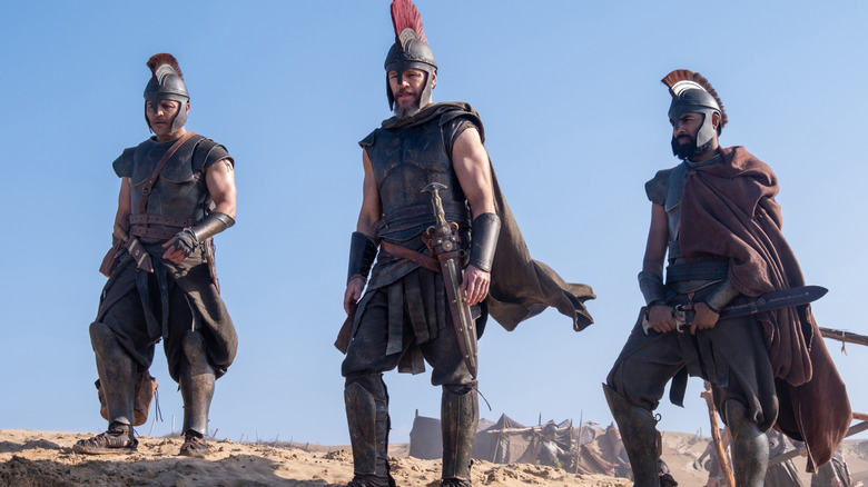 Matt Damon as Odysseus dressed in Greek armor standing on a hill next to Jimmy Gonzales as Cepheus and Himesh Patel as Eurylochus in The Odyssey