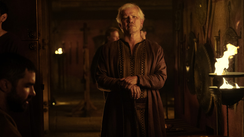 John Leguizamo as Eumaeus leaning on a cane and standing inside a hall lit by firelight in The Odyssey