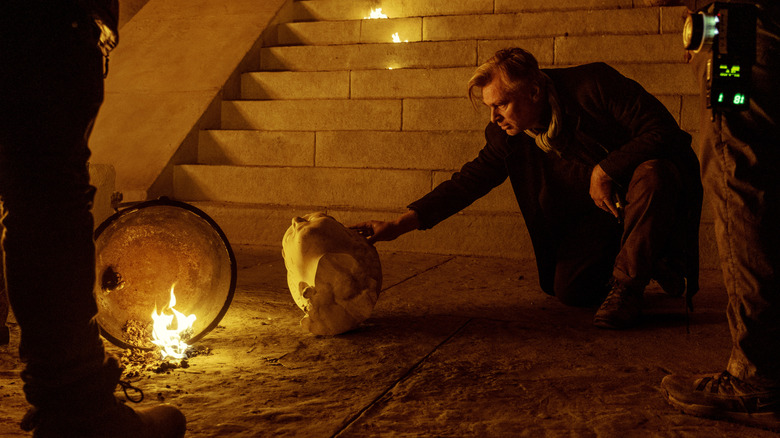 Director Christopher Nolan crouching by a staircase and moving a broken statue into place surrounded by crew on the set of The Odyssey