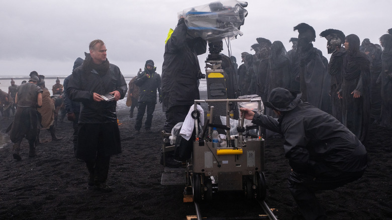 Director Christopher Nolan wearing a rain jacket and standing next to a camera on a beach in front of several extras on the set of The Odyssey