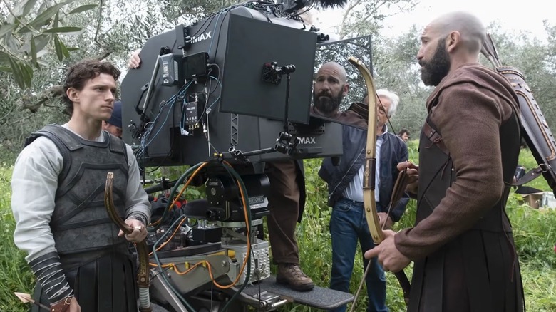 Tom Holland as Telemachus holding a bow and standing next to an IMAX camera next to Jon Bernthal as Menelaus on the set of The Odyssey