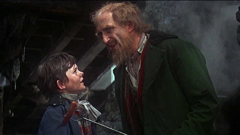 Fagin grins while holding a sausage on a stick in front of Dodger in Oliver!