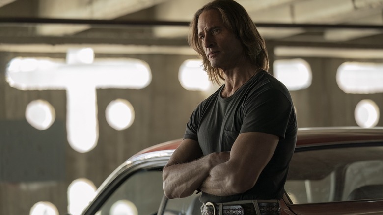 Josh Holloway as Jim Ellis stands with his arms crossed and relying on a red car to Duster