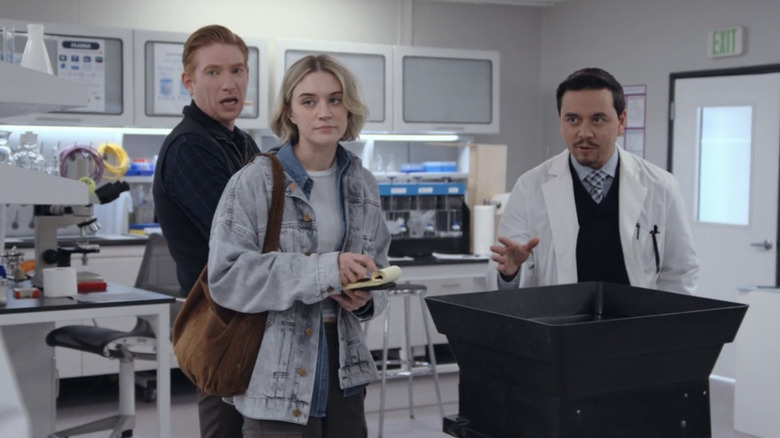 Domnhall Gleeson as Ned Sampson and Chelsea Frei as Mare Pritti stand in a research facility alongside a man in a lab coat in The Paper
