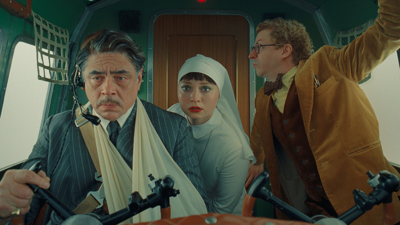 Benicio del Toro as Zsa-zsa Korda flying a plane with his arm in a sling with Mia Threapleton as Liesl and Michael Cera as Bjorn looking concerned in The Phoenician Scheme