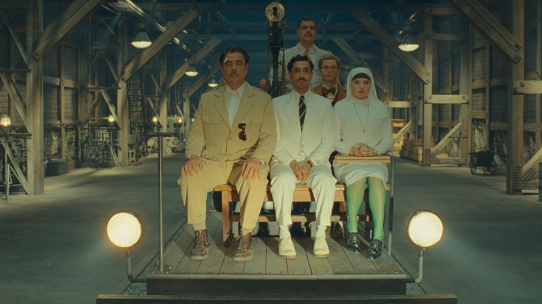 Benicio del Toro as Zsa-zsa Korda, Riz Ahmed as Prince Farouk, Mia Threapleton as Liesl, and Michael Cera as Bjorn seated in a warehouse in The Phoenician Scheme
