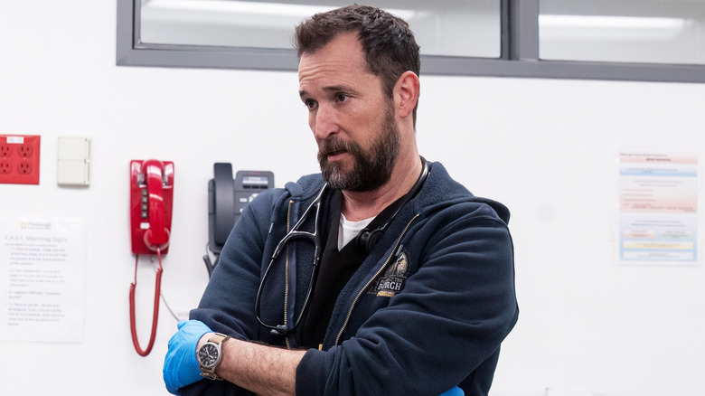 Noah Wyle's Dr Michael Robinavitch stands with his arms folded in a hospital room in The Pitt