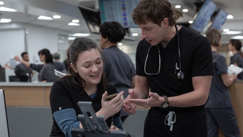 Dr. Santos holds Dr. Whitaker's phone and smiles at what's on the screen, while Whitaker stands nearby looking at her in The Pitt