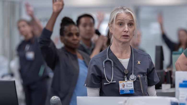 Nurse Dana has a tired and surprised expression on her face while multiple hospital employees raise their hands behind her on The Pitt