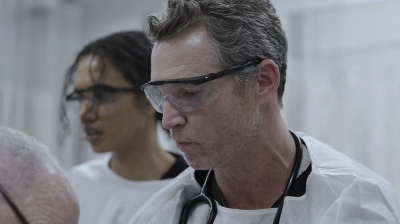 Supriya Ganesh as Dr. Mohan stands behind Sean Hatosy as Dr. Abott in a surgery room in The Pitt