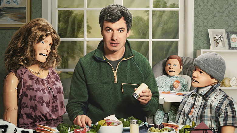 nathan fielder sitting at a kitchen table next to women and two children dolls in the show the rehearsal