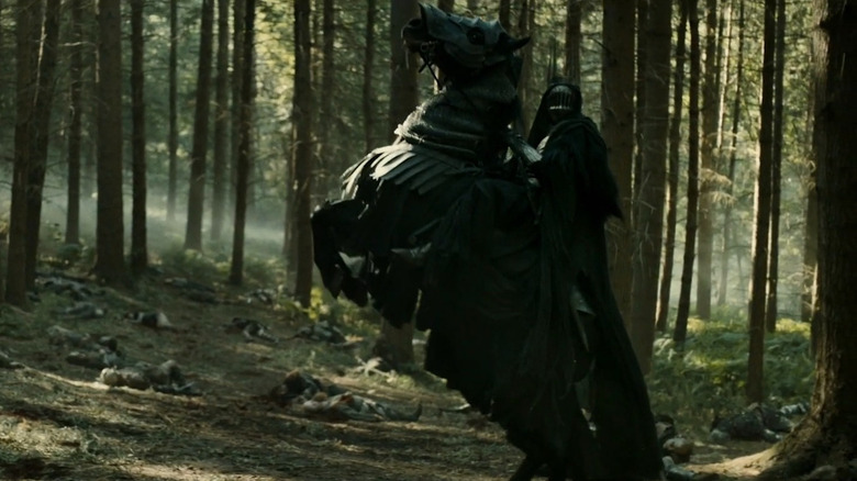 Zubin Varla as Khamûl dressed in black and riding a horse in a forest littered with dead bodies in The Lord of the Rings: The Rings of Power