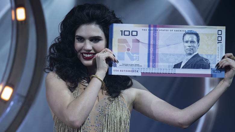 A woman holding a giant New Dollar bill featuring the face of Arnold Schwarzenegger in 2025's The Running Man