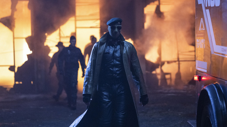Lee Pace as Evan McCone wearing a mask and trench coat standing in front of a burning building in The Running Man