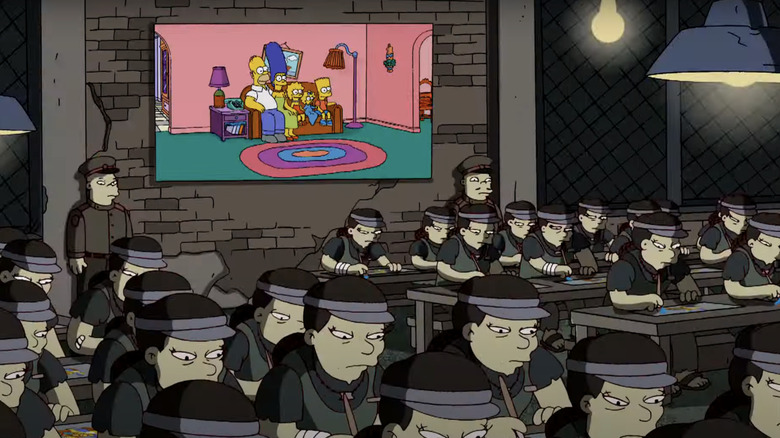 The Simpsons sit on a couch in the background of a sweat shop where animators work on desks in The Simpsons