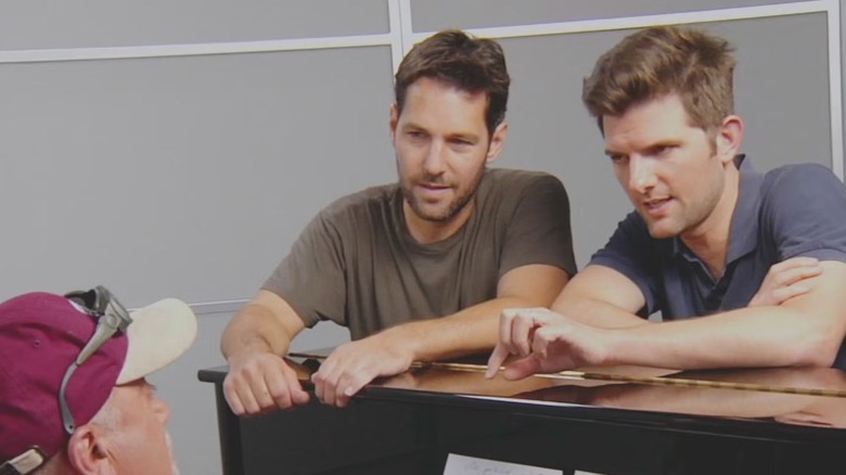 Billy Joel sits at a piano, looking up at Paul Rudd and Adam Scott, who lean over the top of the piano, in The Greatest Event in Television History