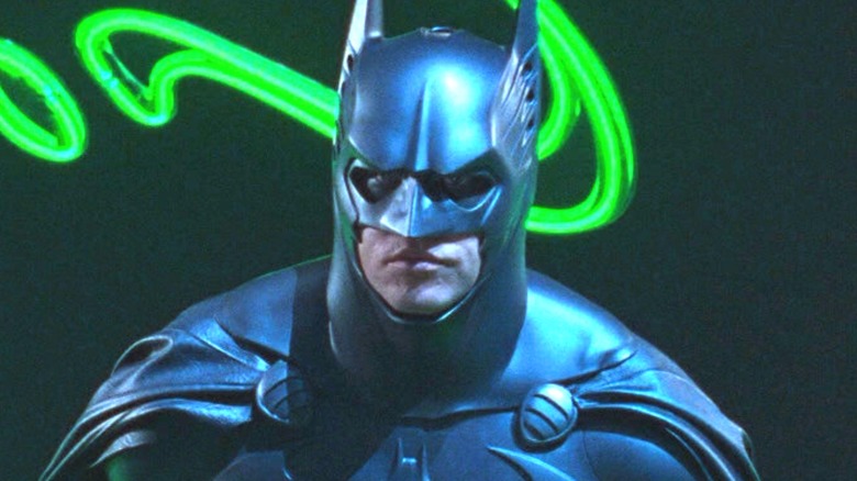 Val Kilmer stands in the Batsuit in front of neon question marks