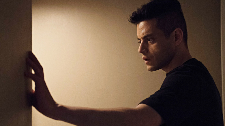 Rami Malek as Elliot Alderson holding arm out against a wall in a drab room in Mr. Robot