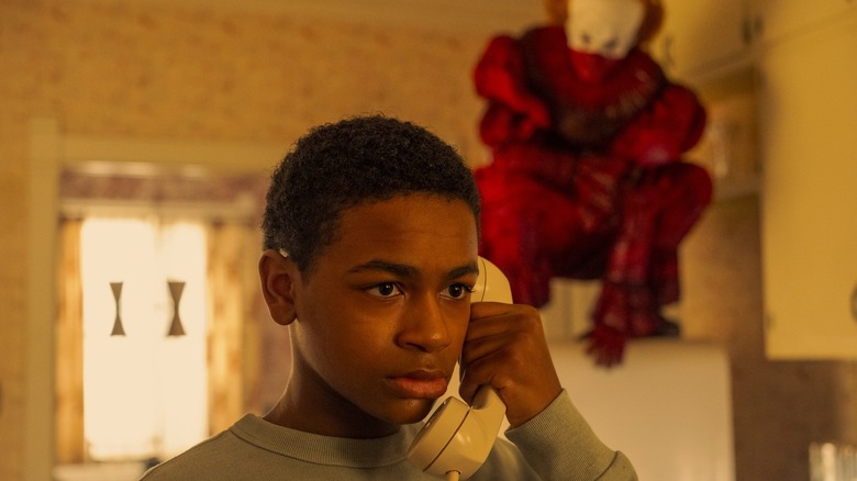 Will Hanlon van Blake Cameron James houdt een telefoon tegen zijn oor terwijl Pennywise van Bill Skarsgård op de achtergrond hurkt in It Welcome to Derry