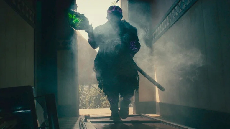 The Toxic Avenger (Peter Dinklage) walks into a hallway covered in smoke and shadow holding a glowing mop in The Toxic Avenger (2023)