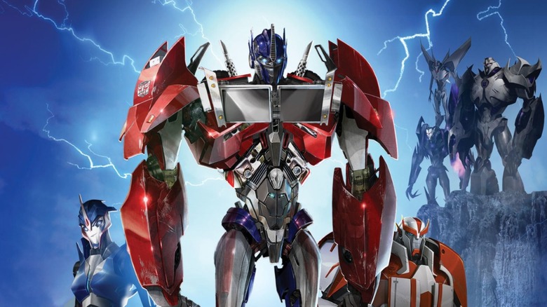 Arcee, Optimus Prime, and Ratchet standing together with Megatron, Starscream, and a Decepticon trooper lurking on a nearby cliff in the Transformers: Prime poster