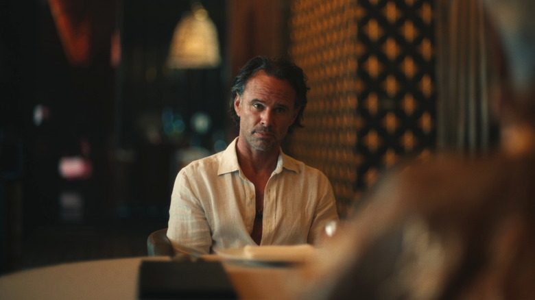 Walton Goggins in the role of Ric, looking skeptical while sitting at a table in a white lotus